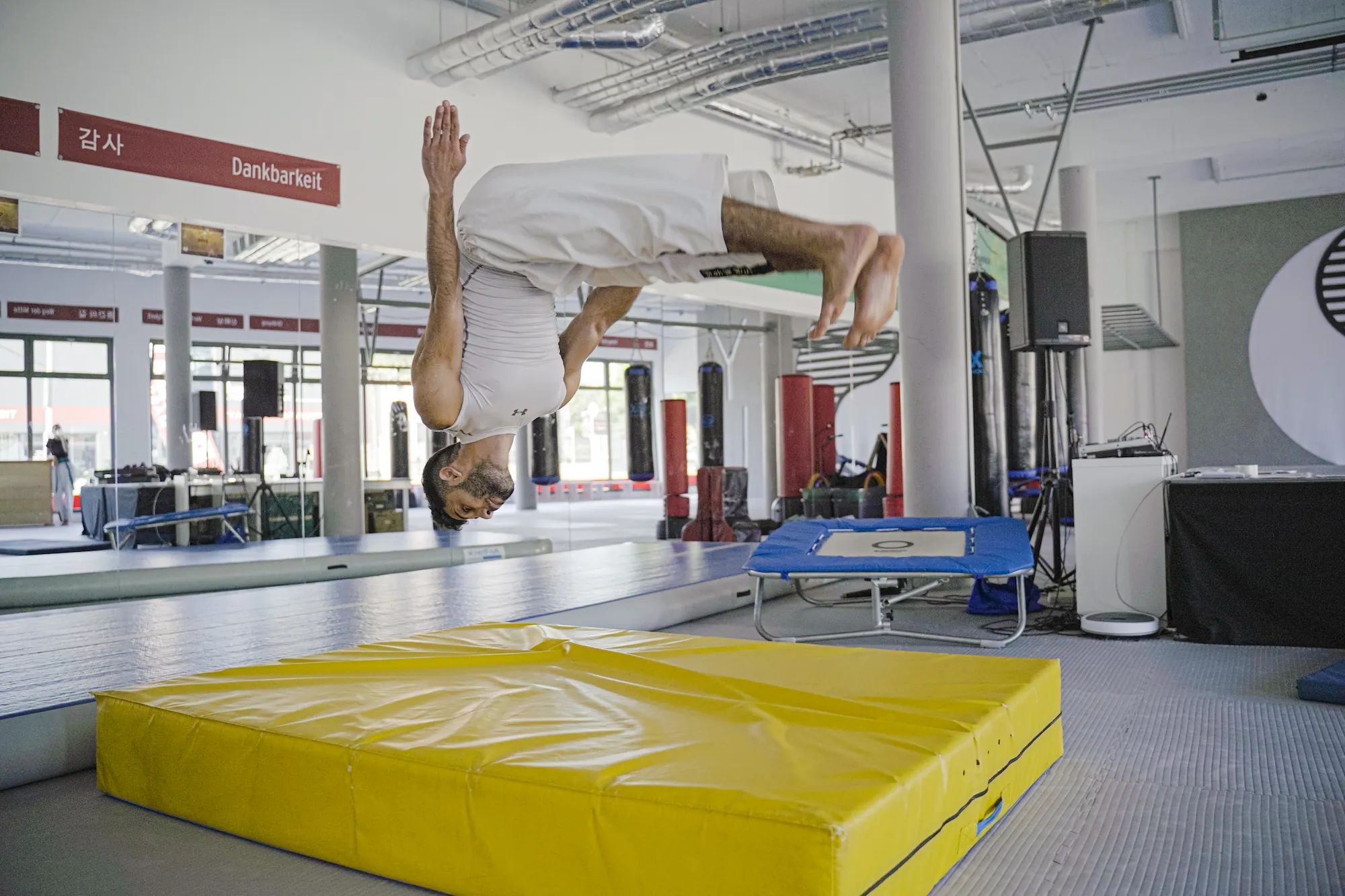 Sportler vollführt einen Rückwärtssalto über einer gelben Matte im Trainingsraum.